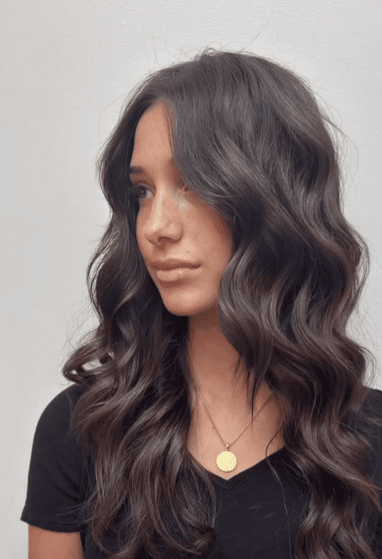 Woman with long, wavy brown hair looking to the side, wearing a black shirt and necklace.