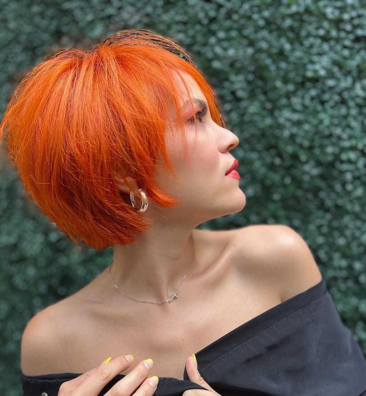 Short-haired woman with bright orange hair, wearing black off-shoulder attire against greenery.